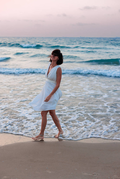 szczęśliwa młoda dziewczyna w białej letniej sukience spaceruje po plaży o zachodzie słońca. dziewczyna taniec na plaży. - Zdjęcie, obraz