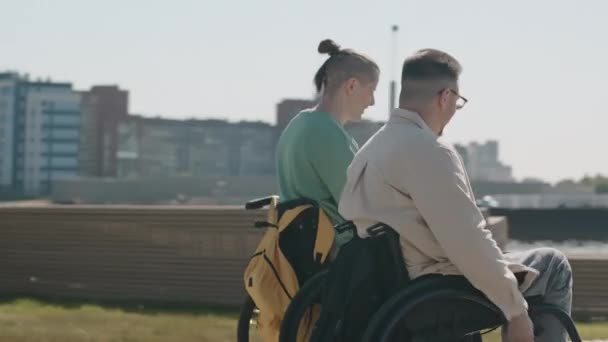 Tracking shot of two cheerful Caucasian male friends racing in wheelchairs on summer city embankment at daytime - Footage, Video