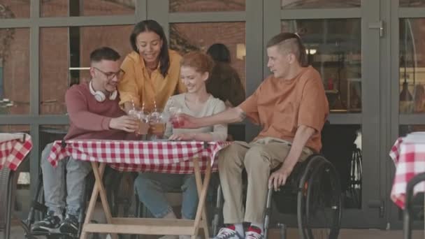 Retrato de duas jovens mulheres e homens em cadeiras de rodas sorrindo para a câmera enquanto aplaudem com óculos de plástico com limonadas enquanto comem juntos no doce café de verão ao ar livre - Filmagem, Vídeo