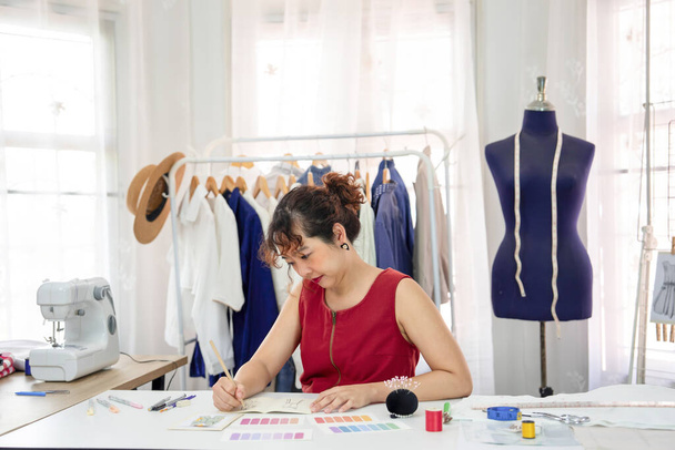 молодая азиатская дизайнер эскиз одежды дизайн на столе, профессиональный портниха работает в ее современной студии моды. design, dressmaking, tailoring, business, industry - Фото, изображение