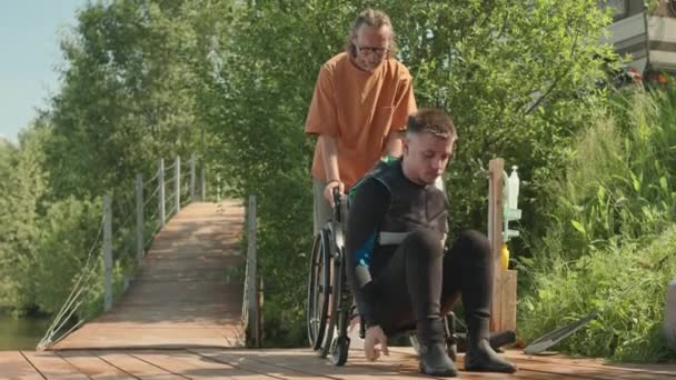 Young Caucasian male surfer in wetsuit getting on wheelchair at wooden pier after surfing in river on sunny day with helpful instructor assisting him - Footage, Video
