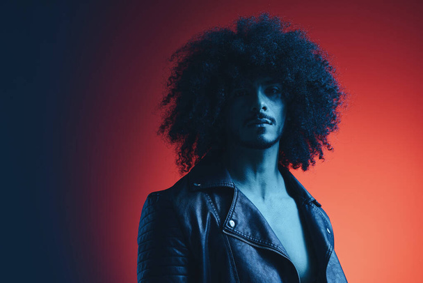 Портрет человека моды с вьющимися волосами на красном фоне многонациональный, цветной свет, черная кожаная куртка тренд, современная концепция. Высокое качество фото - Фото, изображение