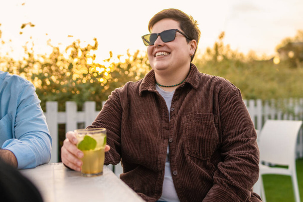 Een niet-binair individu dat geluk uitstraalt, genietend van een sereen moment buiten met een drankje in de hand. Het beeld legt de essentie van authenticiteit en genderdiversiteit vast. - Foto, afbeelding