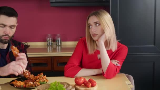 Een jonge brunette eet kippenvleugels in de keuken aan tafel. Een jonge blonde vrouw kijkt er verveeld naar. Eet fastfood. Relaties in het gezin. - Video