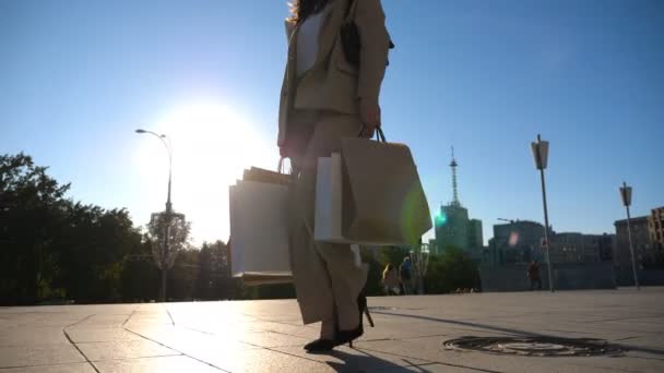 Fashionable lady carries paper packets walking along city square. Young beautiful woman holds shopping bags going at urban street after purchases. Concept of leisure, sales and discounts. Slow motion. - Filmmaterial, Video