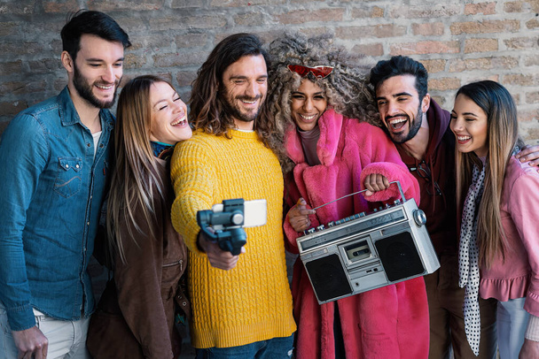 Ryhmä nuoria aikuisia nauraa tiiliseinää vasten, pitäen retro boomboxia ja kuvaten selfie-videota mobiililaitteella. Niiden monipuolinen ulkonäkö ja ilo sekoittavat modernia nuoruutta vintage-viboilla. - Valokuva, kuva