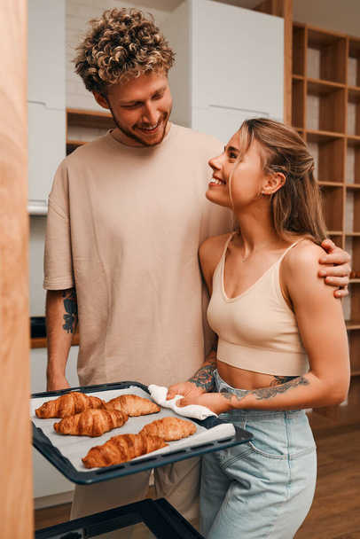 Молодая пара влюбленных печет круассаны на кухне вместе, готовится к завтраку, разговаривает и проводит время романтически вместе. - Фото, изображение