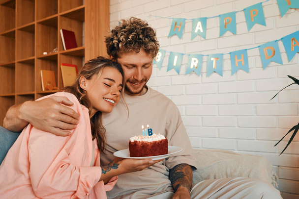 Jong getrouwd stel in liefde vieren hun verjaardag door kaarsen uit te blazen op een taart terwijl ze in de woonkamer zitten in een gezellig huis. - Foto, afbeelding