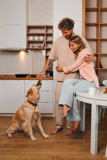 Νεαρό ερωτευμένο παντρεμένο ζευγάρι παίρνει πρωινό το πρωί μαζί στην κουζίνα με το αγαπημένο τους σκυλί σε ένα άνετο σπίτι. - Φωτογραφία, εικόνα
