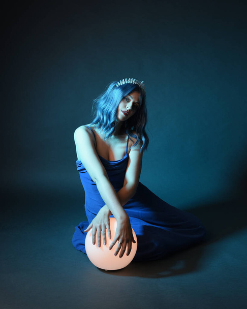 Full length πορτραίτο του όμορφου γυναικείου μοντέλου με μπλε μαλλιά φορώντας λαμπερό φόρεμα μπάλα φαντασίας με Crystal στέμμα, κρατώντας ένα λαμπερό λαμπτήρα σφαίρα. Καθιστή στάση, απομονωμένη σε σκοτεινό φόντο στούντιο - Φωτογραφία, εικόνα