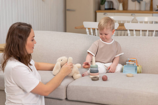 Happy family at home. Mother and baby boy playing with toys in couch at home indoors. Little toddler child and babysitter nanny having fun together. Young woman mom kid son rest in living room - Photo, image
