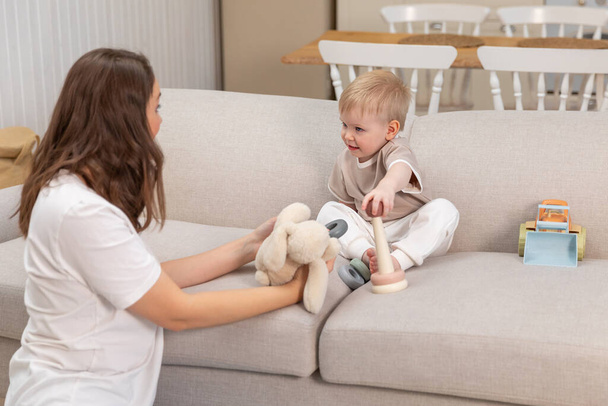 Happy family at home. Mother and baby boy playing with toys in couch at home indoors. Little toddler child and babysitter nanny having fun together. Young woman mom kid son rest in living room - Photo, image