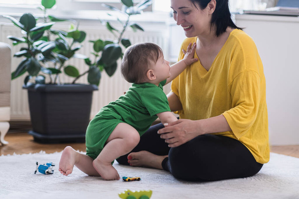 Uroczy chłopiec czołga się do mamusi siedząc na białym dywanie w pobliżu rozrzuconych zabawek szczęśliwa kochająca matka uśmiecha się patrząc na małego syna spędzającego czas w żłobku w domu - Zdjęcie, obraz