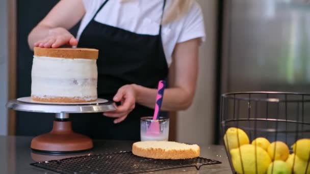 Pastelero profesional en delantal negro que conecta las capas del pastel untadas con la mujer de la crema que hace la pastelería casera de encargo de los productos libres de lactosa que están de pie en la cocina - Imágenes, Vídeo