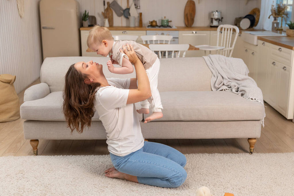 Happy family at home. Mother lifting in air little toddler child son. Mom and baby boy playing having fun together at home. Mother hugs baby with love care. Young mother kid son rest in living room - Photo, Image