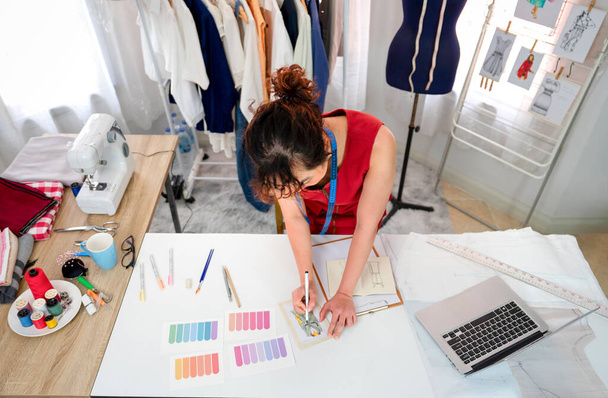 top view fiatal női tervező vázlatos ruházati design az asztalon, egy profi varrónő dolgozik az ő modern divat stúdió. koncepció ruhatervezés, ruhakészítés, szabás, üzlet, ipar - Fotó, kép