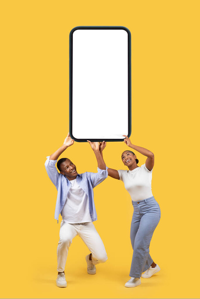 Couple noir tenant un énorme smartphone lourd avec écran noir blanc sur fond de studio jaune, maquette pour l'application mobile ou la publicité sur le site Web, grande offre - Photo, image