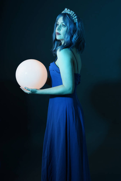 Κοντινό πλάνο πορτρέτο του όμορφου γυναικείου μοντέλου με μπλε μαλλιά φορώντας λαμπερό φόρεμα μπάλα φαντασίας, Crystal στέμμα, κρατώντας λαμπερό λαμπτήρα σφαίρα στην καλλιτεχνική χειρονομιακή στάση. απομονωμένο σε σκοτεινό φόντο στούντιο. - Φωτογραφία, εικόνα