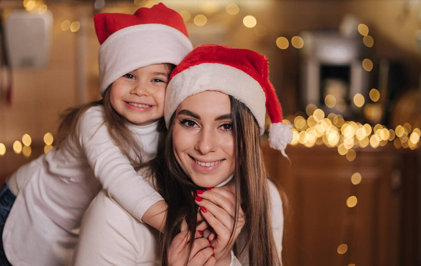Portrait of adorable little daughter with her mom at kitchen during Christmas holidays. Girls in santa hat. Decorative fit tree at the table. Girls make gingerbread. - Photo, Image