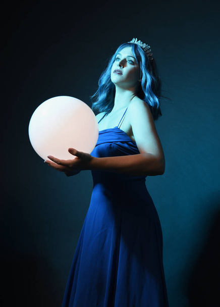 Κοντινό πλάνο πορτρέτο του όμορφου γυναικείου μοντέλου με μπλε μαλλιά φορώντας λαμπερό φόρεμα μπάλα φαντασίας, Crystal στέμμα, κρατώντας λαμπερό λαμπτήρα σφαίρα στην καλλιτεχνική χειρονομιακή στάση. απομονωμένο σε σκοτεινό φόντο στούντιο. - Φωτογραφία, εικόνα