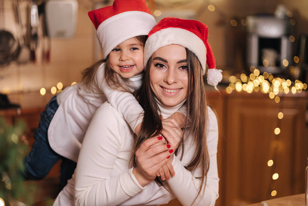 Noel tatilinde annesiyle birlikte mutfakta küçük sevimli bir kızın portresi. Noel Baba şapkalı kızlar. Masadaki dekoratif uygun ağaç. Kızlar zencefilli ekmek yapar.. - Fotoğraf, Görsel