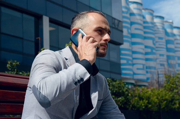 Кавказький дорослий бізнесмен, одягнений в елегантний одяг, сидить на відкритому повітрі серйозно, уважно і продумано, концентрований прослуховування, по телефону, копіювання простору - Фото, зображення