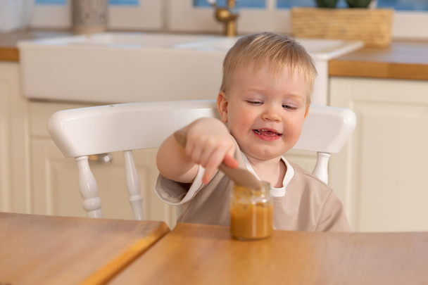 Счастливая семья дома. Мальчик кормит себя на кухне. Маленький мальчик с грязным смешным лицом ест здоровую пищу. Ребенок учится есть самостоятельно, держа ложку. Самопитание - Фото, изображение