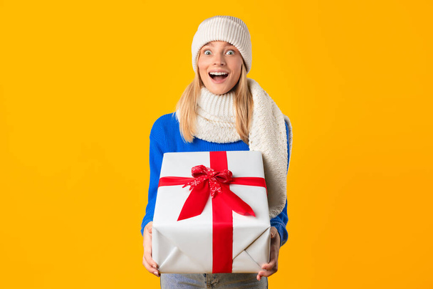 Überglückliche Frau in Winterkleidung klammert sich freudig an großes Weihnachtsgeschenk mit roter Schleife und strahlt Urlaubsstimmung vor gelbem Hintergrund aus - Foto, Bild
