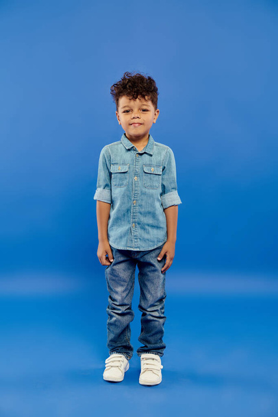mignon et bouclé afro-américain préscolaire garçon en tenue de denim élégant posant sur fond bleu - Photo, image