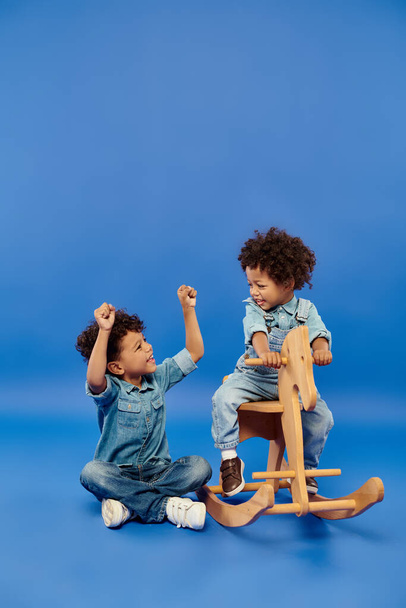happy african american toddler boy in stylish denim clothes sitting on rocking horse near brother - Photo, Image