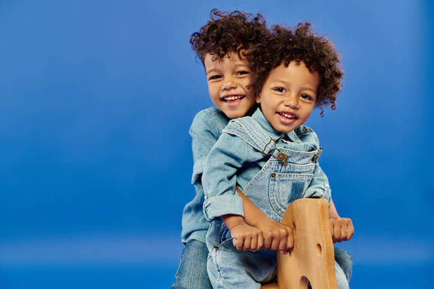 happy african american brothers in stylish denim clothes sitting on rocking horse on blue backdrop - Фото, изображение