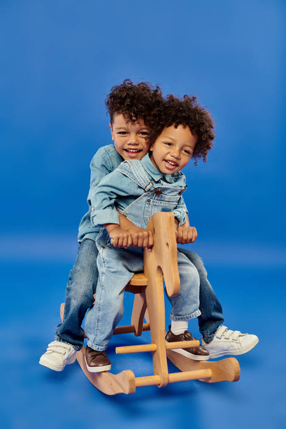 feliz afroamericano chicos en elegante denim ropa sentado en balanceo caballo en azul telón de fondo - Foto, Imagen