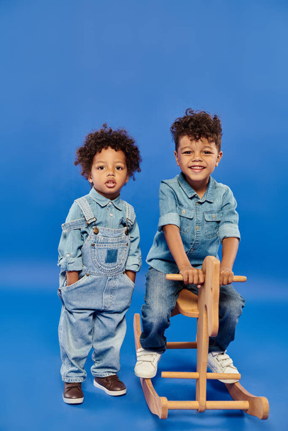 aranyos afro-amerikai csecsemő fiú elegáns farmer ruhák áll közel testvére hintaló - Fotó, kép