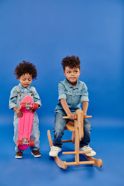 african american boy in stylish denim clothes holding penny board near brother on rocking horse - Photo, Image