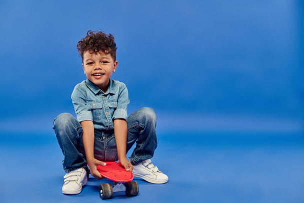 niño preescolar afroamericano lindo en ropa de mezclilla elegante sentado en penny board en azul - Foto, imagen