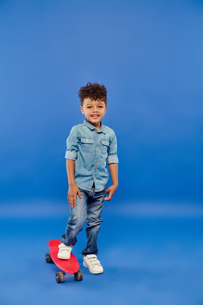 cute african american preschooler boy in stylish denim clothes standing on penny board on blue - Photo, Image