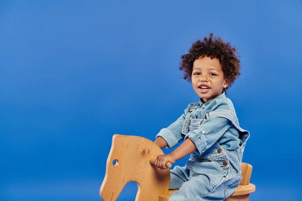 χαρούμενο αφροαμερικανό παιδί με τζιν ρούχα κάθεται σε κουνιστή καρέκλα σε μπλε φόντο - Φωτογραφία, εικόνα