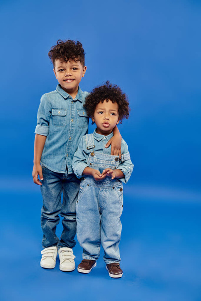 niño preescolar afroamericano feliz en ropa de mezclilla abrazando al hermano pequeño en fondo azul - Foto, Imagen