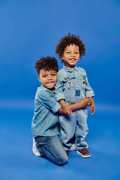 niño preescolar afroamericano feliz en ropa de mezclilla abrazando hermano pequeño en fondo azul - Foto, Imagen