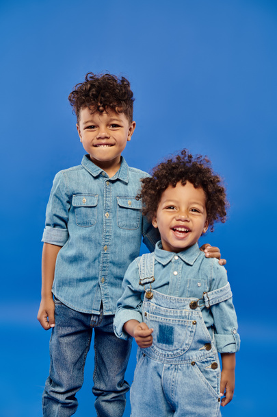 joyeux garçon préscolaire afro-américain en vêtements en denim debout avec frère tout-petit sur bleu - Photo, image