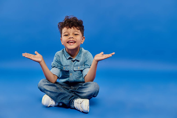 joyeux et frisé afro-américain préscolaire garçon en denim vêtements assis et gestuelle sur bleu - Photo, image