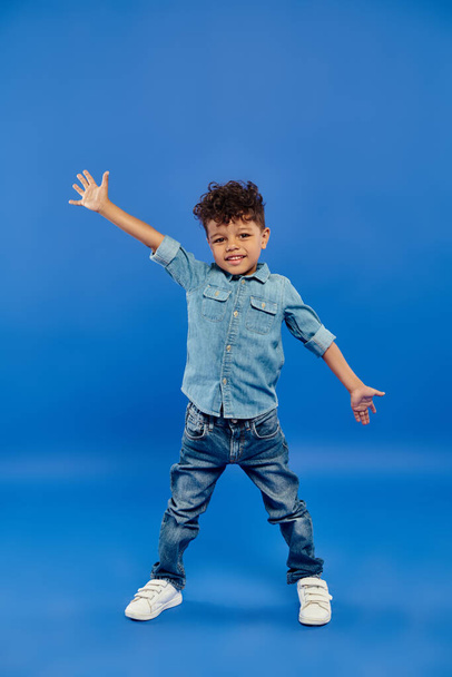 niño preescolar afroamericano alegre y rizado en ropa de mezclilla de pie y gestos en azul - Foto, imagen