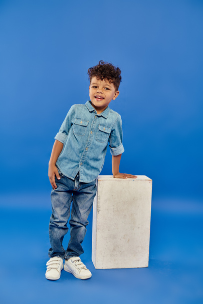 šťastný a kudrnatý africký americký předškolák chlapec v džínových šatech opírající se o bílou kostku na modré - Fotografie, Obrázek