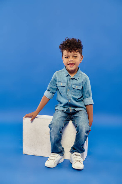 wesoły i kręcony afrykański amerykański przedszkolak chłopiec w dżinsowych ubraniach siedzi na białym sześcianu na niebieskim - Zdjęcie, obraz