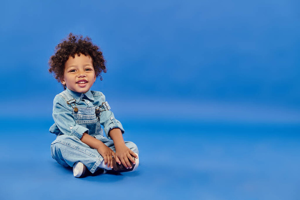 joyful and curly african american toddler boy in denim clothes sitting with crossed legs on blue - Photo, Image