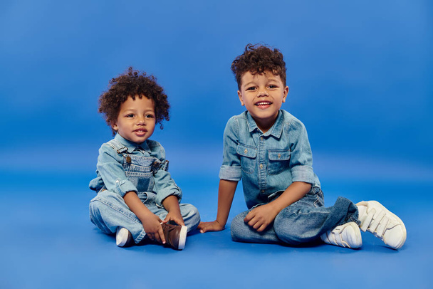 rozkošné africké americké sourozence ve stylovém džínovém oblečení sedí společně na modrém pozadí - Fotografie, Obrázek
