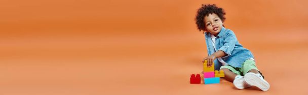 Αφροαμερικάνος νήπιο με denim πουκάμισο κάθεται και παίζει δομικά στοιχεία σε πορτοκαλί, banner - Φωτογραφία, εικόνα