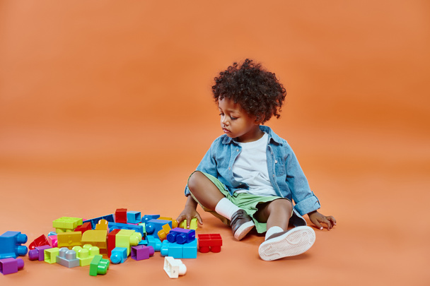 adorable african american toddler boy in denim shirt sitting and playing building blocks on orange - Foto, Imagen