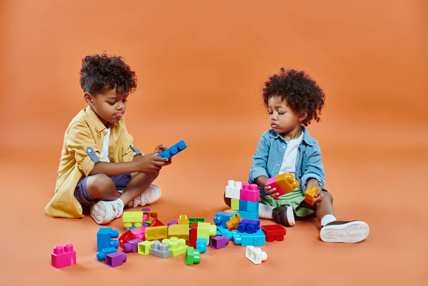 lindo afroamericano hermanos chico en traje casual sentado y jugando bloques de construcción en naranja - Foto, imagen