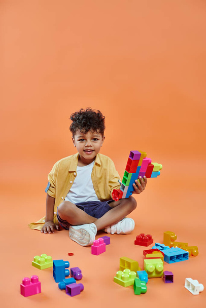 heureux afro-américain garçon en tenue décontractée assis et jouer blocs de construction colorés sur orange - Photo, image